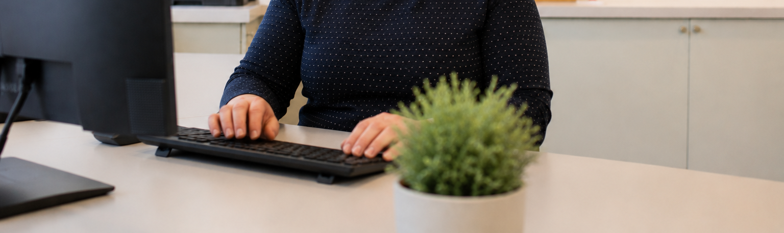 Person tippt an einem Büro-PC, im Vordergrund eine kleine Topfpflanze auf dem Schreibtisch