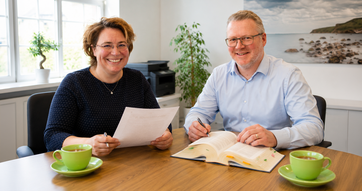 Die Partner – Spieckermann und Goetz Zwei lächelnde Personen sitzen an einem Bürotisch mit Unterlagen und Fachbuch und besprechen gemeinsam ein Dokument
