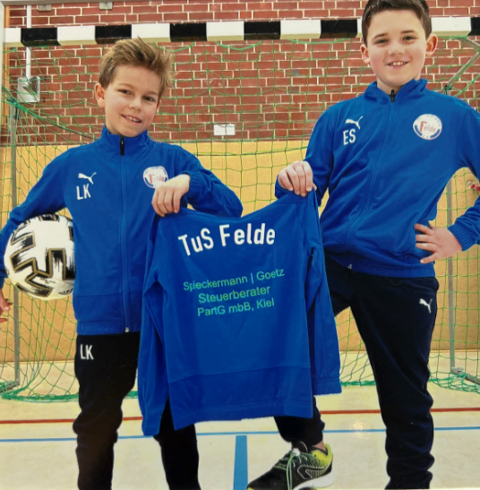 Wir sponsoren den TuS Felde Zwei Kinder in blauen TuS Felde Trainingsanzügen halten ein Trikot mit Vereinslogo und Sponsorenaufdruck Spieckermann Goetz Steuerberater PartG mbB Kiel